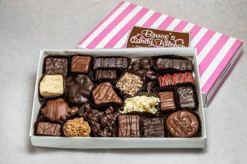 A box of assorted chocolates, featuring various shapes and toppings, set against a pink and white striped background.