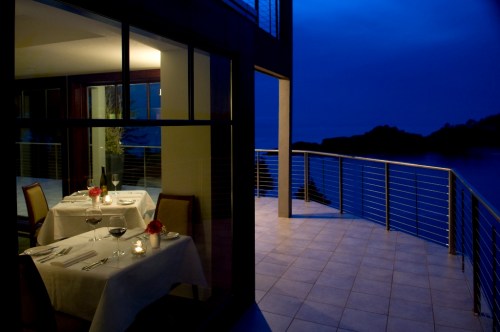 A romantic dining setup with two tables, candles, and flowers, overlooking a serene waterfront at dusk.