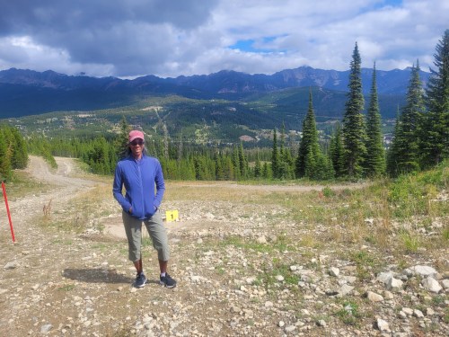 A person stands on a gravel path surrounded by trees and mountains under a partly cloudy sky.