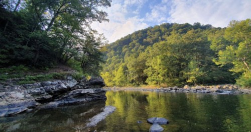 A serene river scene with lush green trees and a rocky shore, reflecting the sky and surrounding hills.