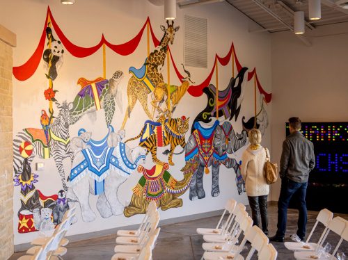 A colorful mural of circus animals, including giraffes and elephants, with two people admiring it in a spacious room.