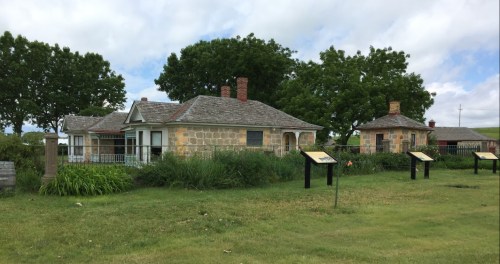 Historic stone houses surrounded by greenery and informational signs under a partly cloudy sky.