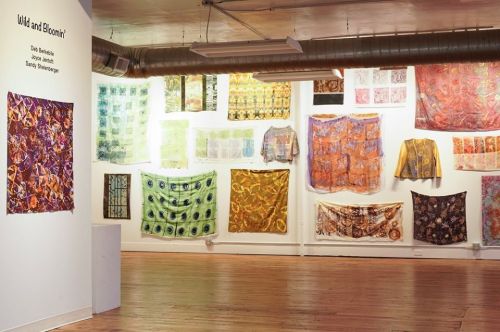 Art gallery wall displaying colorful textile artworks titled "Wild and Bloomin'" by various artists.