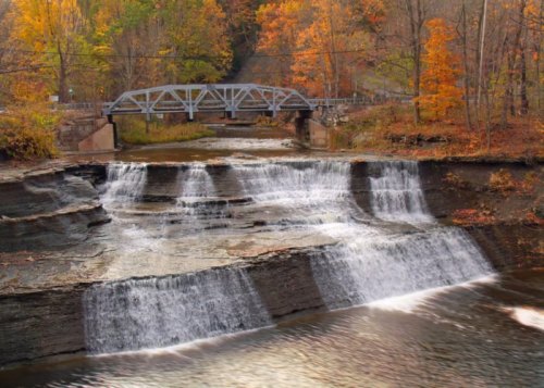 A serene waterfall cascades over rocky ledges, surrounded by autumn foliage and a bridge in the background.