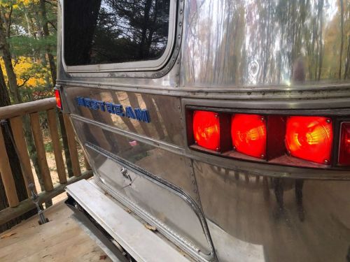 A shiny Airstream trailer with red taillights, parked near a wooden deck surrounded by autumn trees.