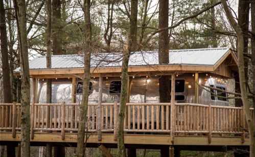 A treehouse with a wooden deck, nestled among trees, featuring a metal exterior and string lights.