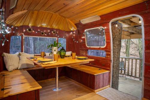 Cozy interior of a rustic cabin with wooden decor, a dining table, and string lights, surrounded by nature.