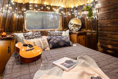 Cozy interior of a rustic cabin with a bed, guitar, and decorative pillows, illuminated by string lights.