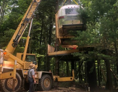 A crane lifts an Airstream trailer above a wooden platform in a forested area. A person watches from below.
