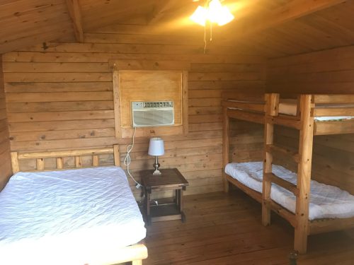 Cozy wooden cabin interior with two beds, a nightstand, and an air conditioning unit on the wall.