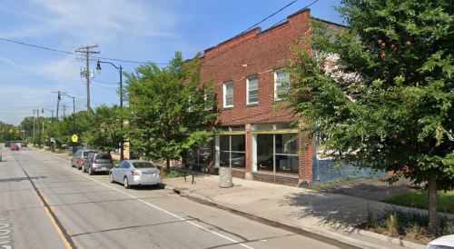 A street view of a brick building with large windows, lined with trees and parked cars along the road.