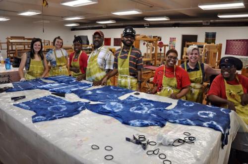 A group of eight people in aprons smiles around a table with blue tie-dye fabric and supplies in a workshop setting.