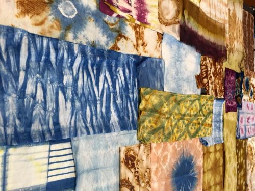 A colorful array of patterned fabrics laid out, showcasing various tie-dye and shibori techniques in blues, browns, and yellows.