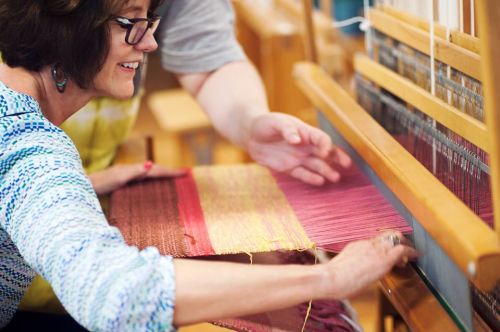 A woman smiles as she weaves fabric on a loom, assisted by another person.