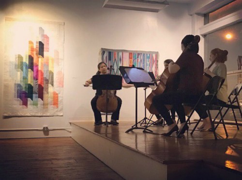 A string quartet performs on a small stage in an art gallery, with colorful artwork displayed on the walls.