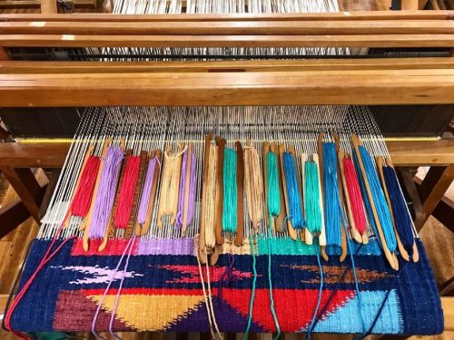 A wooden loom with colorful threads and shuttles, showcasing a vibrant woven textile in progress.