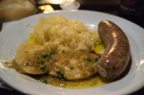 A plate of sauerkraut, fish fillet in sauce, and a sausage, served with a roll in the background.