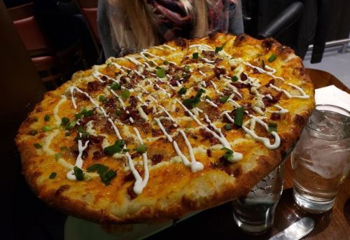A large pizza topped with cheese, bacon, green onions, and drizzled with sauce, served on a wooden table.