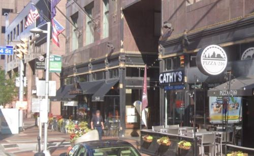 Street view of a city block featuring restaurants, outdoor seating, and flags.