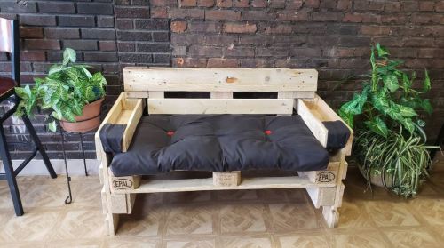 A rustic wooden pallet sofa with a black cushion, surrounded by potted plants against a brick wall.