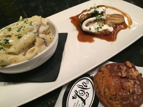 A plate featuring creamy pasta, a meat dish with sauce, and a cookie topped with bacon bits on a dark surface.