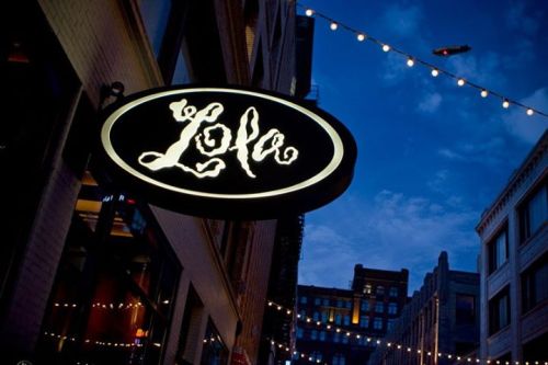 A glowing sign for "Lola" hangs above a street, with buildings and a twilight sky in the background.