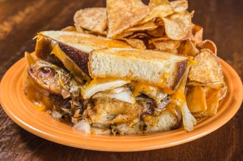 A hearty sandwich with melted cheese, grilled onions, and crispy chips on the side, served on an orange plate.