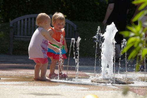 Two young children play joyfully in a splash fountain on a sunny day, surrounded by water jets and greenery.