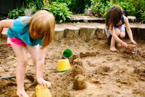 Two children play in a sandy area, building sandcastles with buckets and tools, surrounded by greenery.