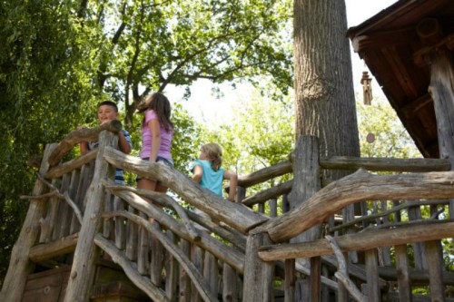 Three children play on a wooden structure surrounded by trees, enjoying a sunny day outdoors.