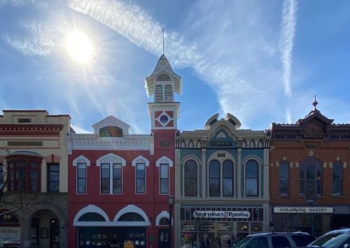 Historic buildings line a street under a bright sun and wispy clouds, showcasing unique architectural styles.