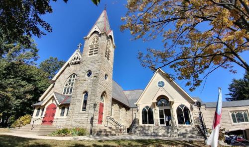 Historic stone church with a tall steeple, surrounded by trees and a clear blue sky.