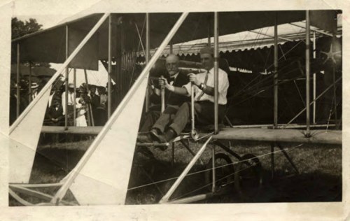 Two men seated in an early airplane, smiling and holding instruments, with a crowd and tents in the background.
