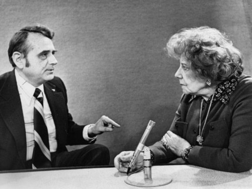 A man and woman engage in a serious conversation at a table, with a small object between them. Black and white photo.