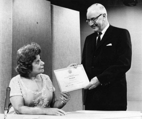 A man presents a certificate to a woman, both smiling, in a studio setting with a microphone on the table.