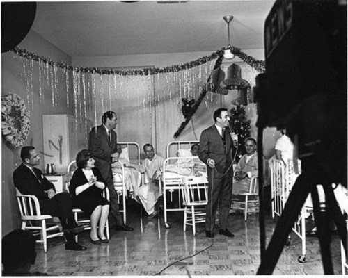A black-and-white photo of a group in a room with hospital beds, festive decorations, and a man speaking to the audience.