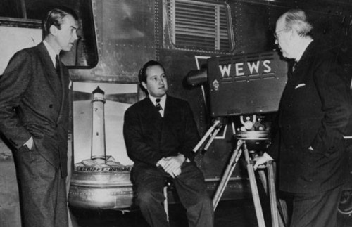 Three men in suits discuss in front of an old television camera labeled "WEWS," with a vintage bus in the background.