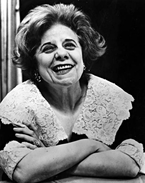 A smiling woman with curly hair and a lace collar, resting her arms on a table, exuding warmth and joy.