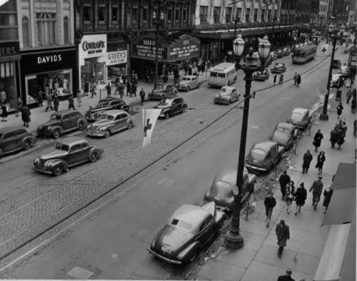 A busy city street in black and white, featuring vintage cars, pedestrians, and storefronts from the mid-20th century.