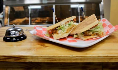 Two sandwiches with shredded chicken and lettuce on a plate, served on a wooden table next to a small bell.