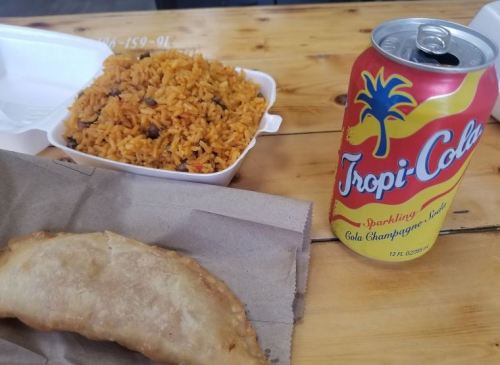 A plate of yellow rice with beans, a fried pastry, and a can of Tropi-Cola on a wooden table.