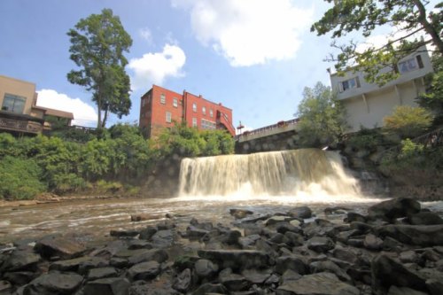 A waterfall cascades over rocks, surrounded by greenery and buildings under a blue sky with fluffy clouds.
