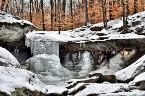 A frozen waterfall surrounded by snow-covered rocks and trees with autumn leaves in a serene winter landscape.
