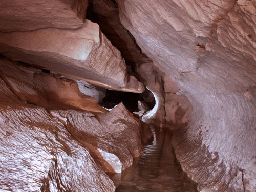A winding cave passage with smooth, illuminated rock formations and a shallow water pool reflecting the light.