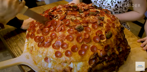 A large, round pizza topped with pepperoni slices, being sliced by a person at a wooden table.