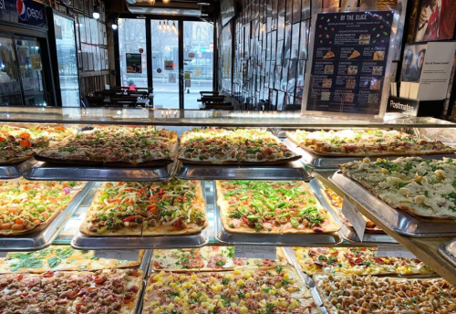 A display of various colorful pizzas in a restaurant, with a view of the street through large windows.