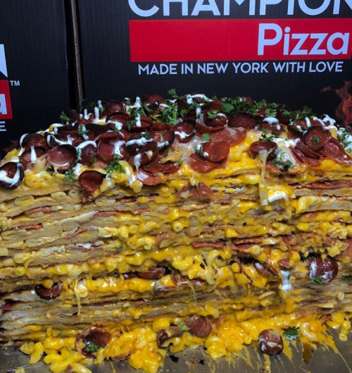 A towering slice of pizza layered with macaroni, cheese, pepperoni, and topped with herbs, served against pizza boxes.
