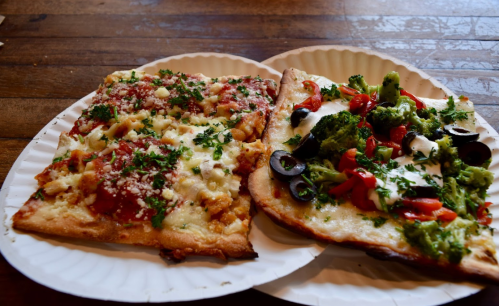 Two slices of pizza on paper plates: one with marinara, cheese, and herbs; the other topped with broccoli, olives, and tomatoes.