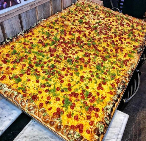 A large rectangular pizza topped with cheese, cherry tomatoes, and green herbs, served on a wooden table.