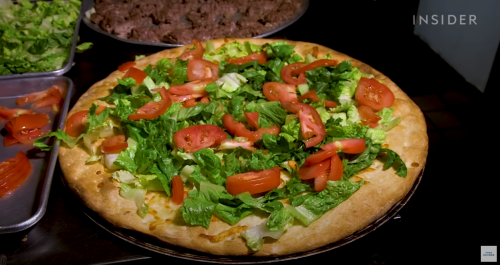 A freshly made pizza topped with lettuce and tomato, with a tray of cooked meat in the background.
